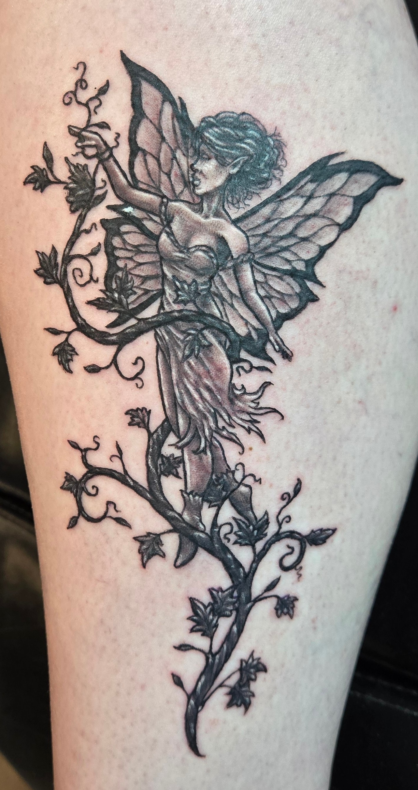 Black and gray custom tattoo of a fairy surrounded by winding vines and leaves
